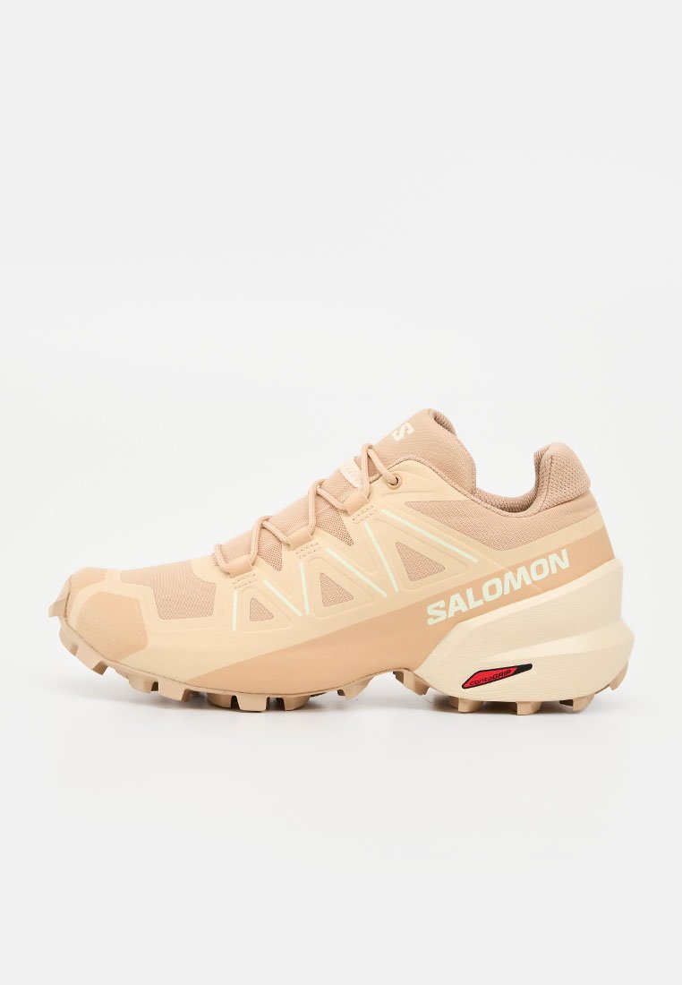 Beiger Salomon Trailrunning-Schuh mit robuster Sohle, reflektierenden Details und gebrandetem Logo an der Seite.