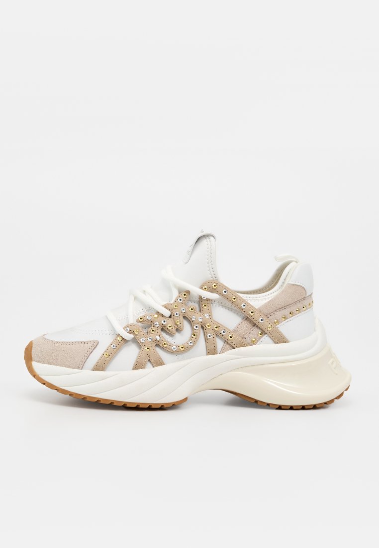 Weißer und beiger Sneaker mit dicker Sohle, verziert mit kleinen goldenen Nieten und Strasssteinen, von der rechten Seite auf einfarbigem Hintergrund gezeigt.