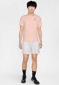 Helles rosa T-Shirt mit kurzen Ärmeln und kleinem Logo, kombiniert mit hellgrauen Sportshorts mit seitlichen Taschen. Schwarze Sportschuhe und Socken.