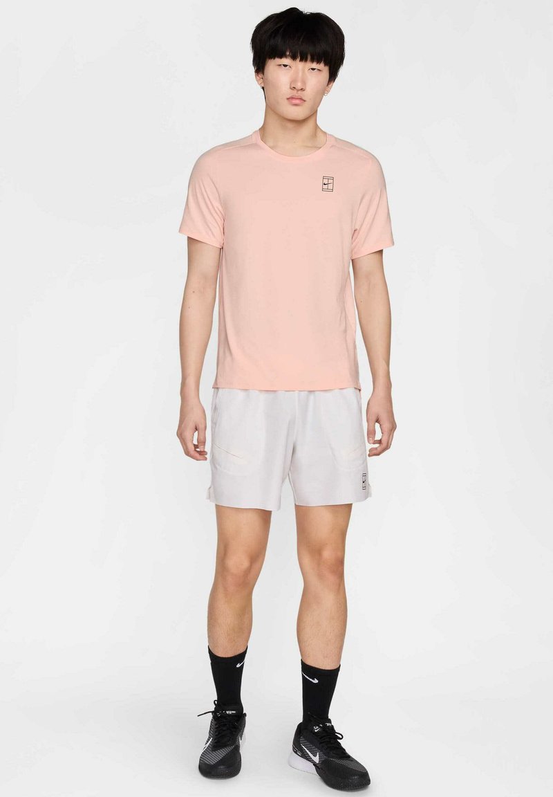 Helles rosa T-Shirt mit kurzen Ärmeln und kleinem Logo, kombiniert mit hellgrauen Sportshorts mit seitlichen Taschen. Schwarze Sportschuhe und Socken.
