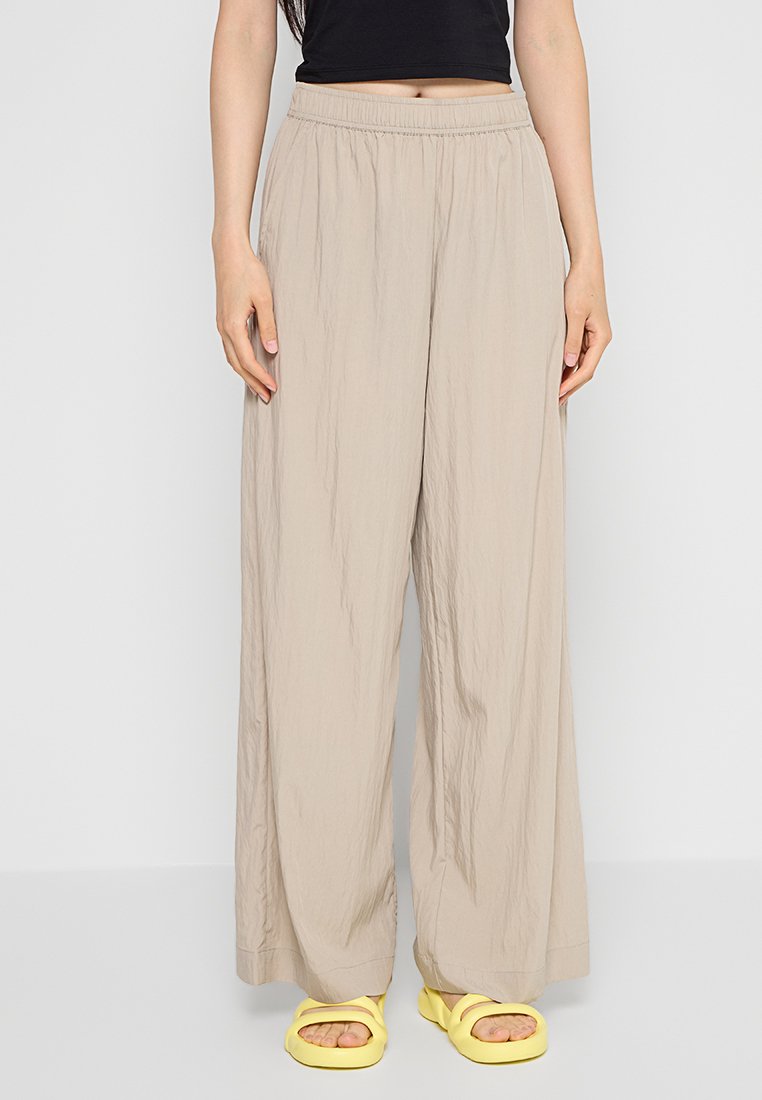 Beige weite Hose mit einem elastischen Bund, aus leichtem, plissiertem Stoff gefertigt. Kombiniert mit gelben Sandalen.
