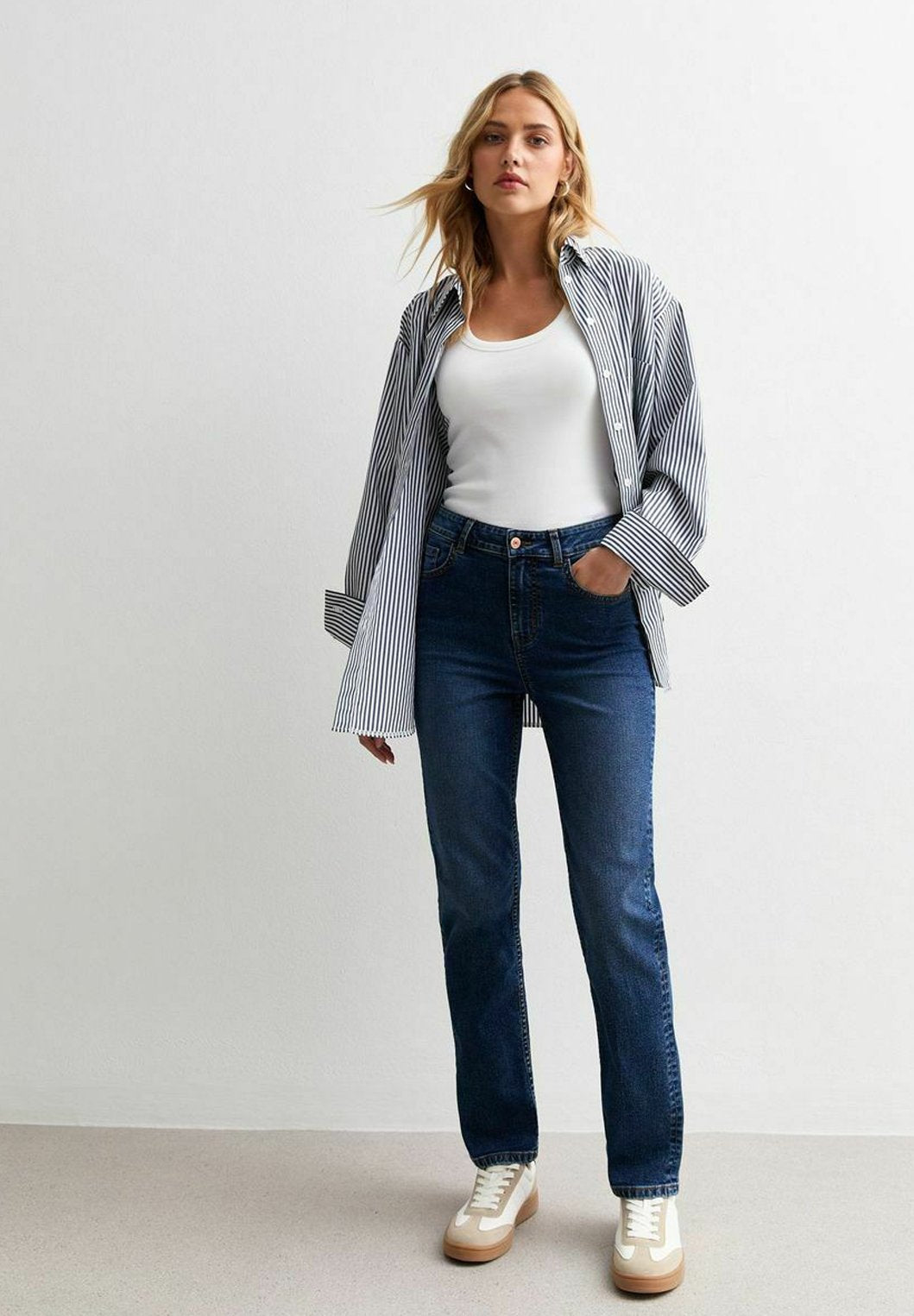 Gestreifte Hemdbluse in Weiß und Blau, getragen über einem weißen Tanktop, kombiniert mit dunklen Jeans und beigen Sneakern.