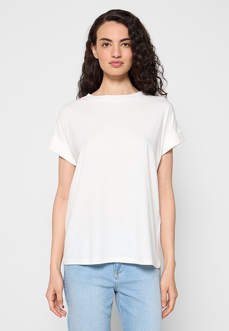 Frau mit langen lockigen Haaren trägt ein schlichtes weißes T-Shirt mit kurzen Ärmeln und hellblaue Jeans, steht vor einem schlichten hellgrauen Hintergrund.