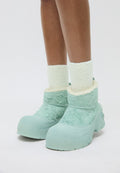 Beine, die dicke, weiße, kuschelige Socken und klobige, hellblaue Winterstiefel mit strukturiertem Stoff und Gummisohlen tragen, vor einem weißen Hintergrund.