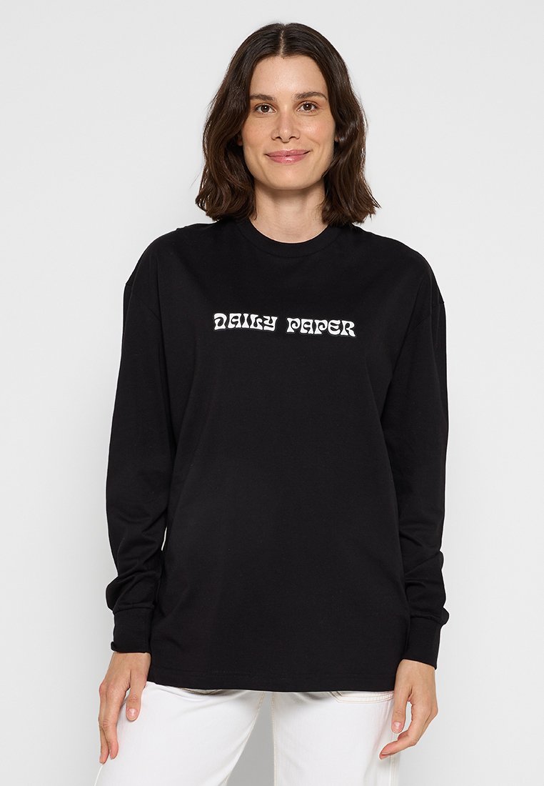 Frau mit schulterlangem dunklem Haar trägt ein schwarzes Langarmshirt mit weißem "DAILY PAPER"-Text, kombiniert mit weißen Hosen, vor hellem Hintergrund.