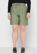 Frau in olivgrünen Zugband-Shorts, schwarzem Langarmshirt und dunkelbraunen Slippern, stehend vor einem schlichten hellen Hintergrund.