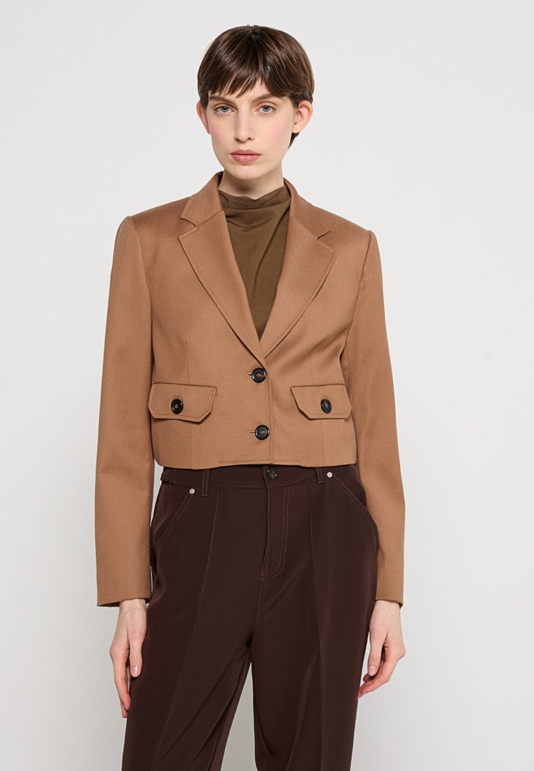 Brauner, kurz geschnittener Blazer mit zwei Fronttaschen, einreihigem Design und schwarzen Knöpfen, kombiniert mit einem braunen Rollkragenpullover und dunkler Hose.