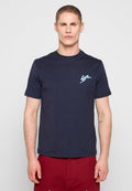 Mann mit kurzen Haaren, der ein marineblaues T-Shirt mit einem kleinen hellblauen Hasenlogo auf der Brust und roten Shorts trägt, steht vor einem einfarbigen Hintergrund.