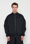 Mann trägt schwarze Jacke mit Reißverschluss und passende Hose mit subtil strukturierten Zahlenmustern, steht vor einfachem weißem Hintergrund.