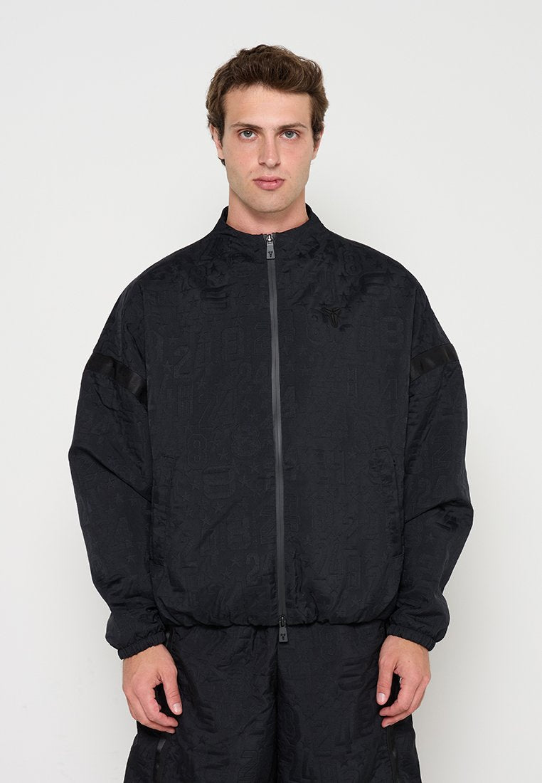 Mann trägt schwarze Jacke mit Reißverschluss und passende Hose mit subtil strukturierten Zahlenmustern, steht vor einfachem weißem Hintergrund.