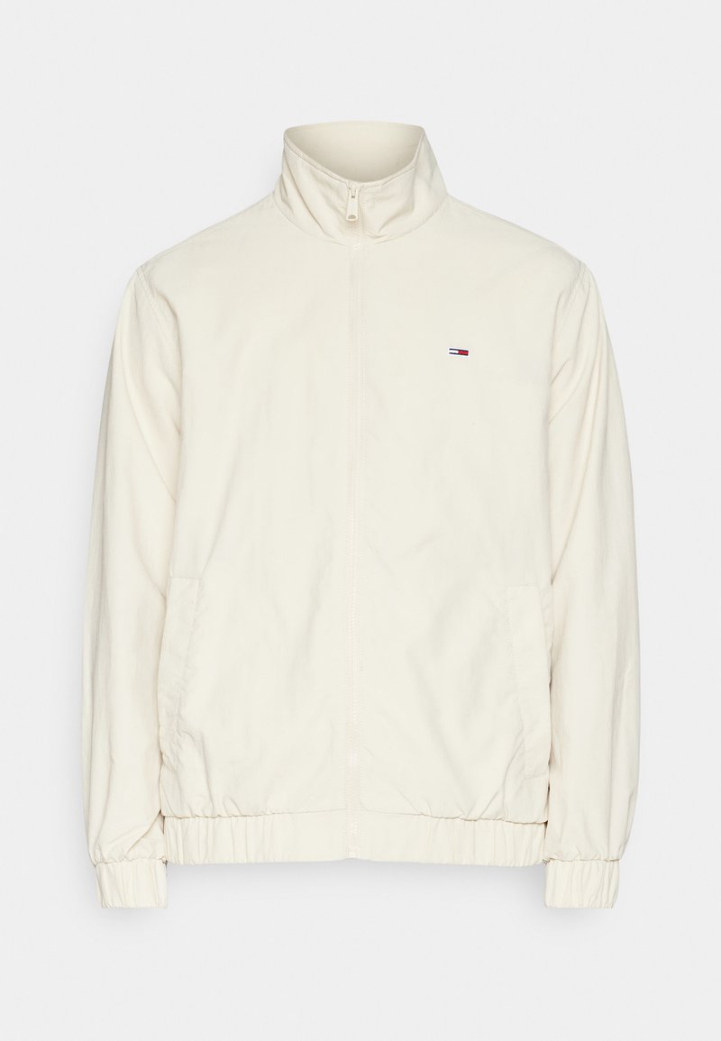 Cremfarbener Reißverschlussjacke mit hohem Kragen, elastischen Bündchen und Saum, mit einem kleinen Logo auf der linken Brust. Glatte Stoffoberfläche.