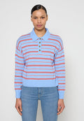 Frau mit dunklen Haaren, die ein hellblaues und rot gestreiftes langärmeliges Poloshirt und blaue Jeans trägt, stehend vor einem schlichten Hintergrund.