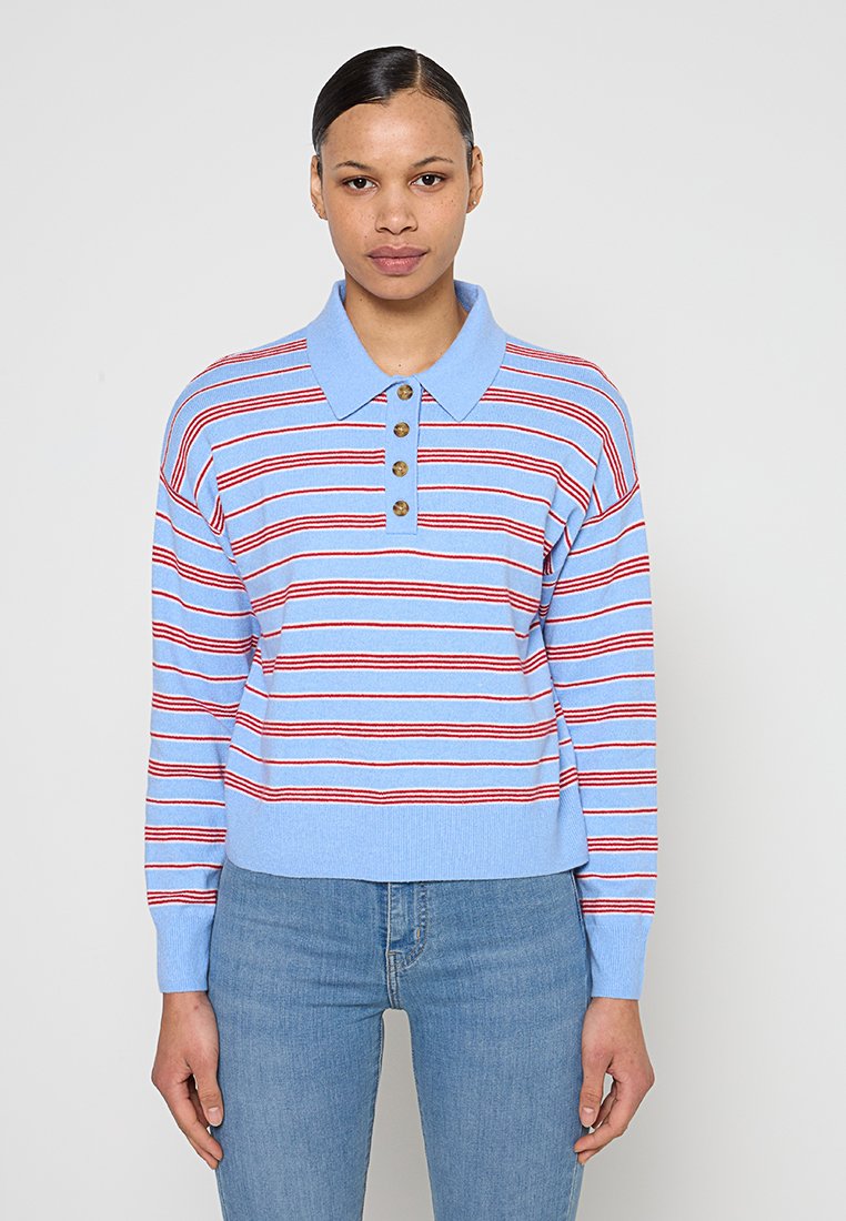 Frau mit dunklen Haaren, die ein hellblaues und rot gestreiftes langärmeliges Poloshirt und blaue Jeans trägt, stehend vor einem schlichten Hintergrund.