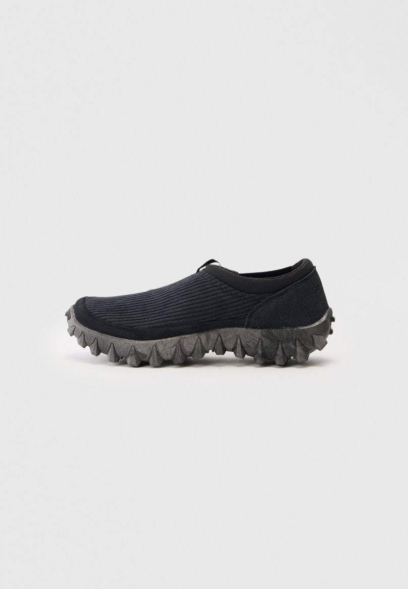 Schwarzer Slipper mit Obermaterial aus geripptem Stoff und robuster, grauer Sohle mit Spitzen, seitlich auf einem schlichten Hintergrund dargestellt.