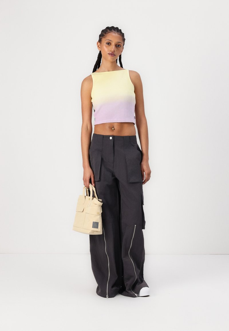 Junge Person mit geflochtenem Haar, trägt ein ärmelloses Crop-Top in Pastellgelb und -lila, weite schwarze Cargohosen mit Reißverschlüssen, hält eine beige Tragetasche.