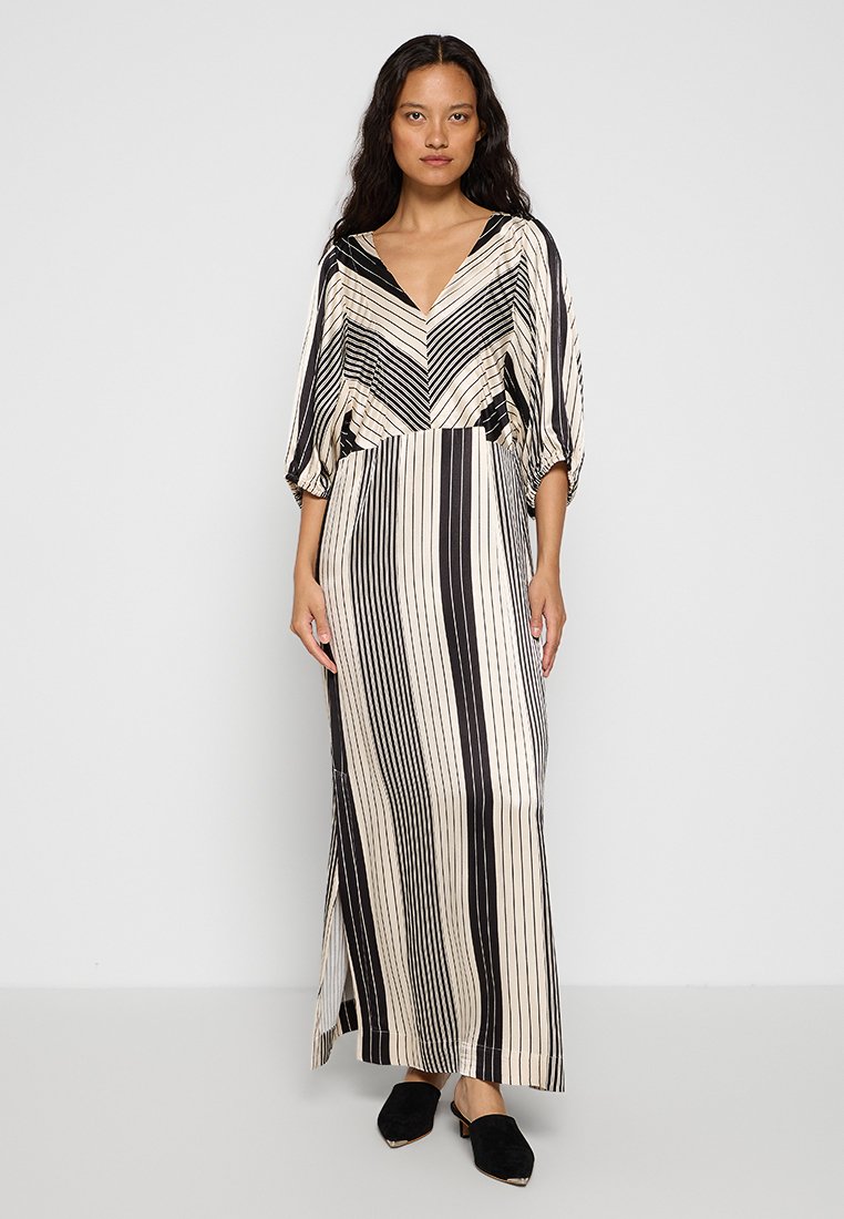 Maxikleid in Creme mit schwarzen vertikalen Streifen. V-Ausschnitt, weite Ärmel und seitliche Schlitze. Weicher Stoff, leicht lockere Passform. Schwarze Schuhe getragen.