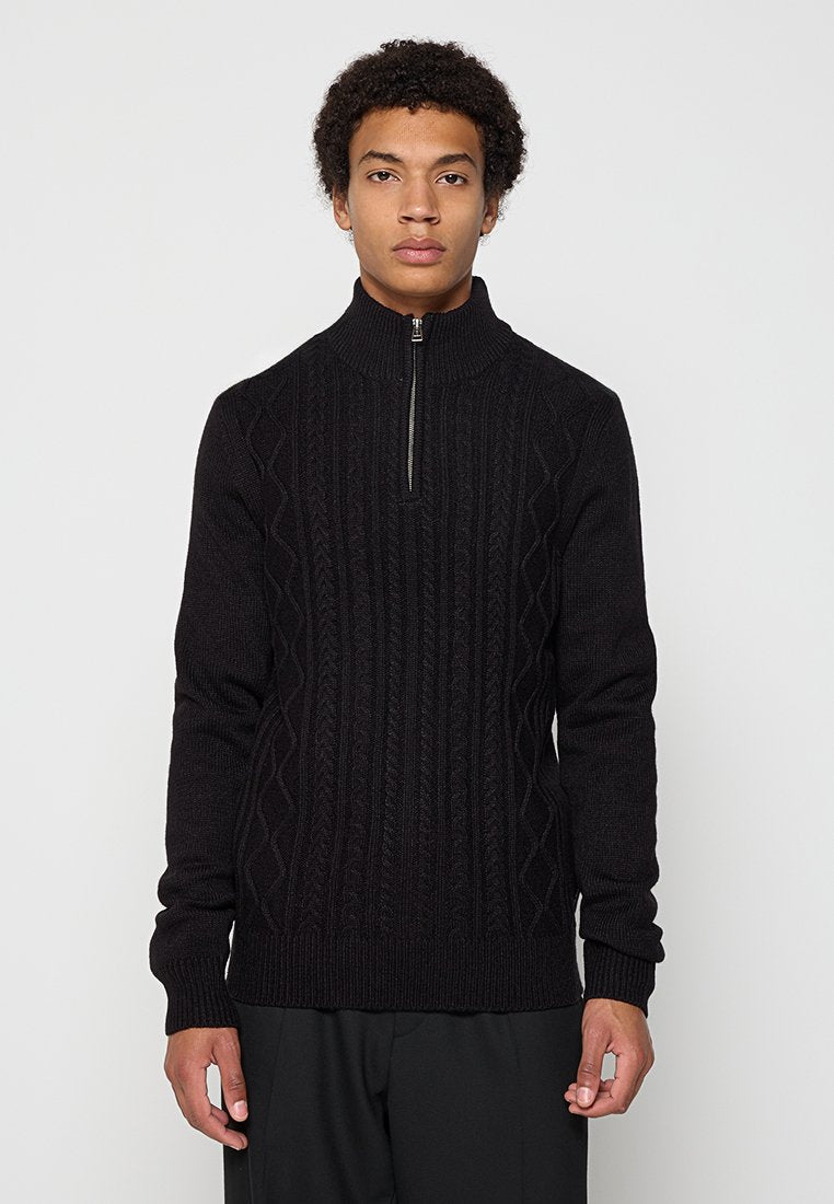 Junger Mann mit lockigem Haar, der einen schwarzen Strickpullover mit einem Viertel-Reißverschluss und schwarze Hosen trägt und vor einem einfarbigen Hintergrund steht.