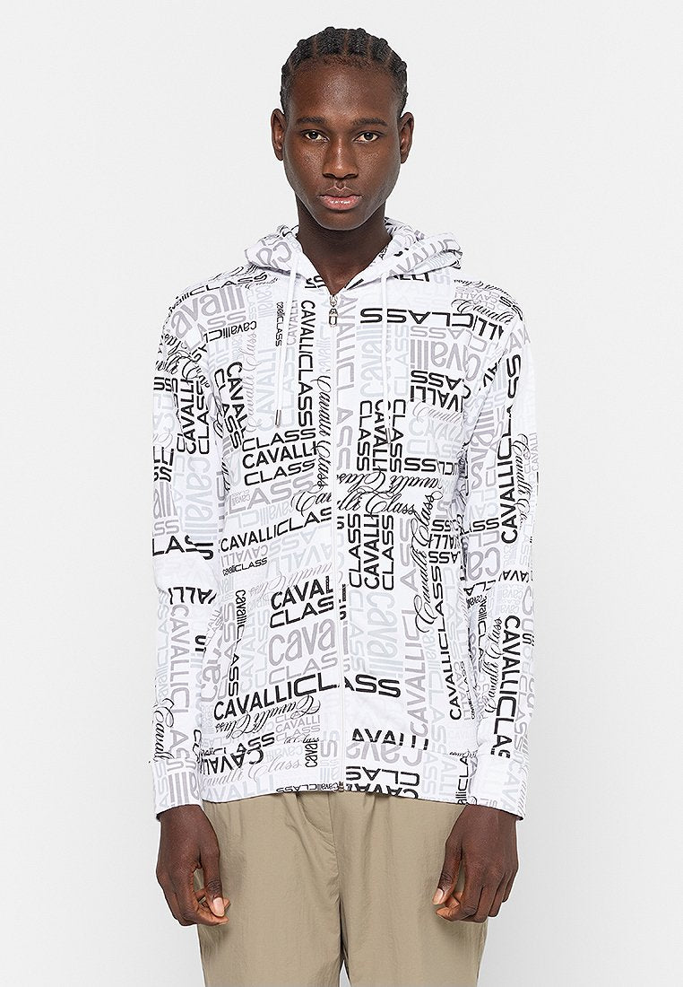 Mann in weißem Reißverschluss-Hoodie mit wiederholtem schwarzen und grauen "CAVALLI CLASS"-Druck, kombiniert mit beigen Hosen, stehend vor einem weißen Hintergrund.