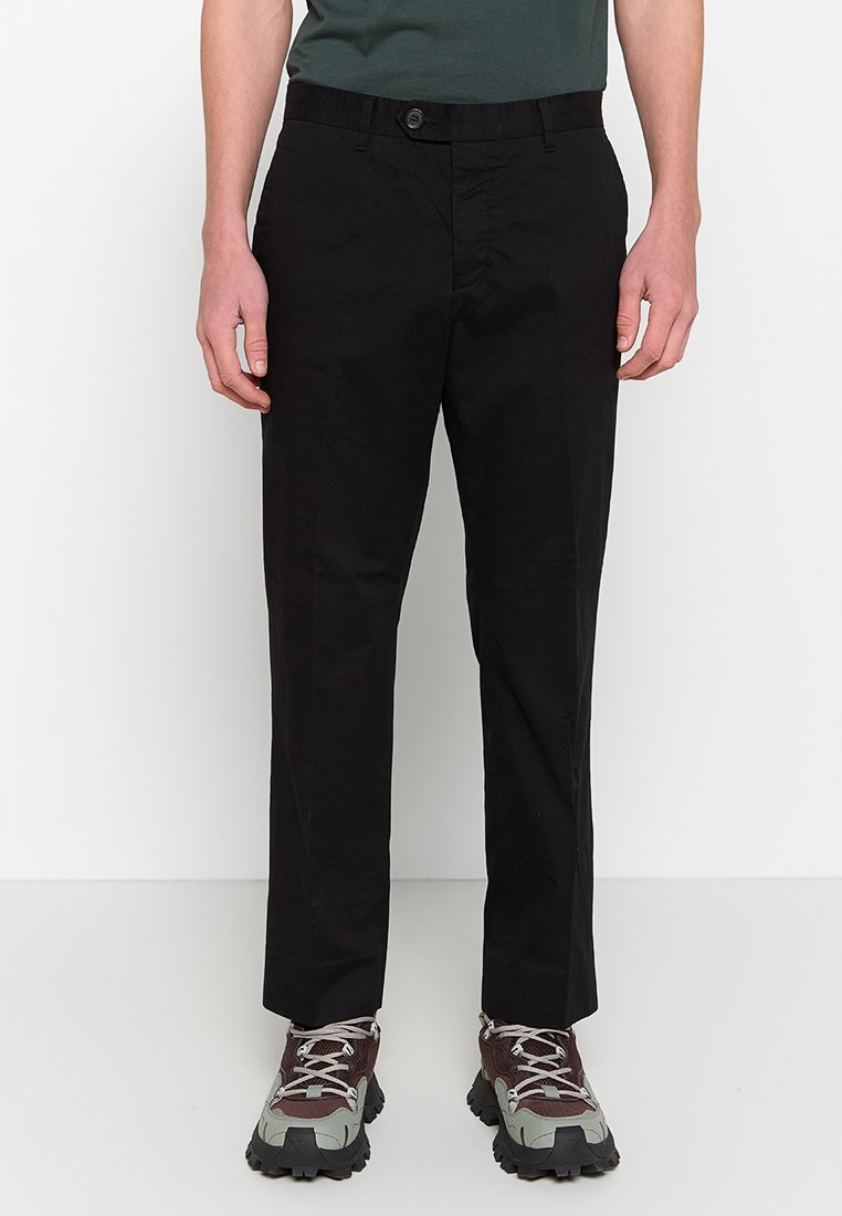 Schwarze Baumwollhosen mit geradem Schnitt, ausgestattet mit einem Knopfverschluss und sichtbaren Taschen. Strukturierter Stoff, kombiniert mit robustem Schuhwerk.