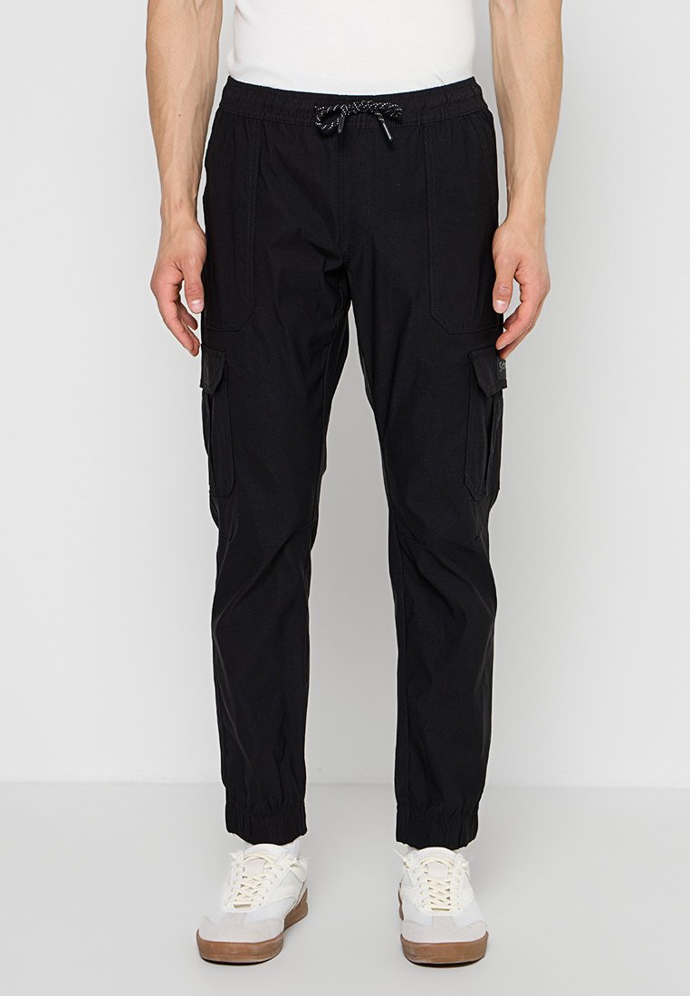 Mann trägt schwarze Cargo-Jogger-Hose mit Kordelzug in der Taille und elastischen Bündchen, kombiniert mit weißen Sneakern.