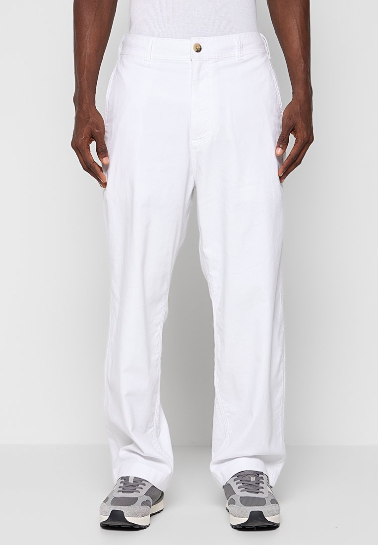 Mann in weißen Hosen und grau-weißen Sneakers, der vor einem schlichten hellen Hintergrund steht, mit entspannten Händen an den Seiten.
