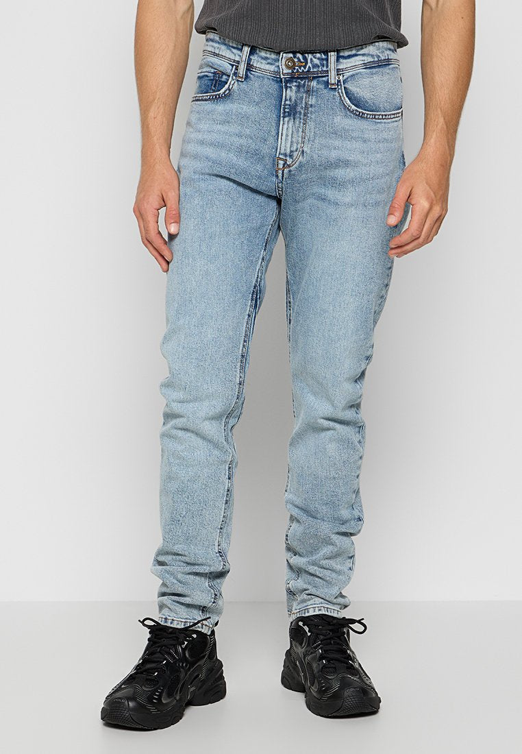 Mann trägt hellblaue Slim-Fit-Jeans und schwarze Schnürschuhe und steht vor einem schlichten weißen Hintergrund.