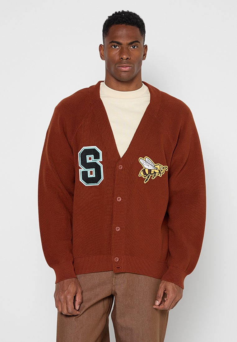Mann trägt eine rostfarbene, geknöpfte Strickjacke mit einem großen "S" und einem gelben Bienen-Patch über einem weißen Hemd und braunen Hosen.