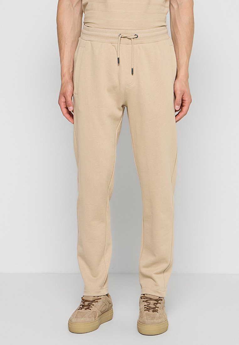 Mann trägt beige Jogginghosen mit Kordelzug in der Taille und beige Turnschuhe, steht vor einem hellgrauen Hintergrund.