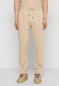 Mann trägt beige Jogginghosen mit Kordelzug in der Taille und beige Turnschuhe, steht vor einem hellgrauen Hintergrund.