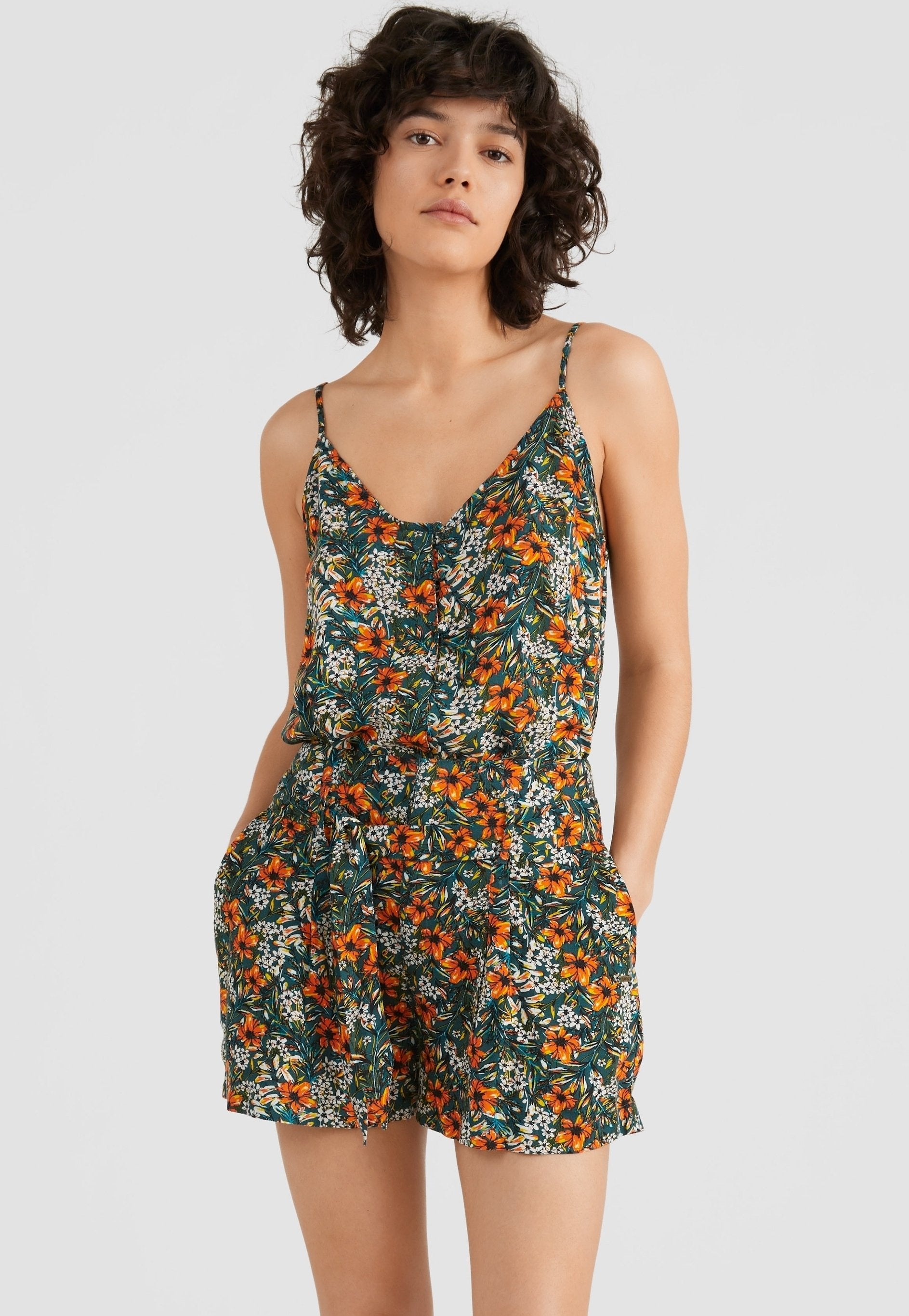 Blümchenbedruckter Spielanzug mit V-Ausschnitt, verstellbaren Trägern und Taschen. Merkmale lebhafte orange-weiße Blumen auf grünem Hintergrund.