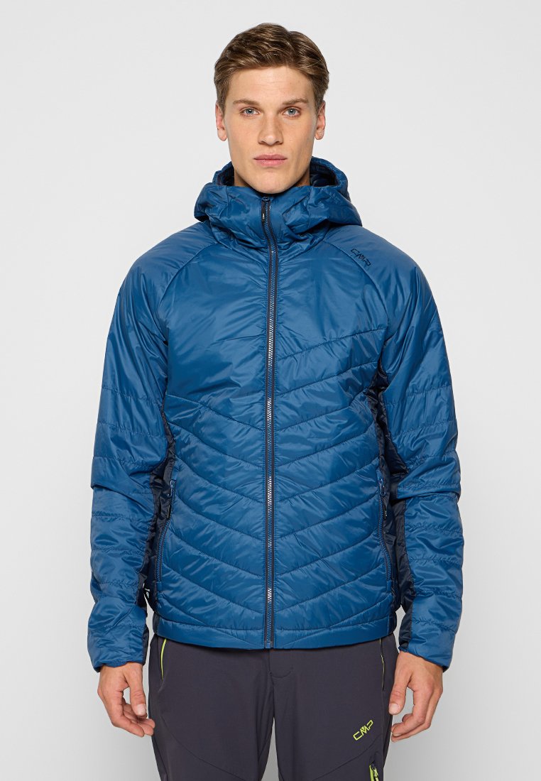 Mann in einer blauen gepolsterten Kapuzenjacke, die zugezippt ist, steht vor einem schlichten hellen Hintergrund.