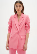 Junge Frau mit langen braunen Haaren, die ein pinkfarbenes, maßgeschneidertes Blazer und passende Hosen trägt, steht vor einem schlichten weißen Hintergrund.