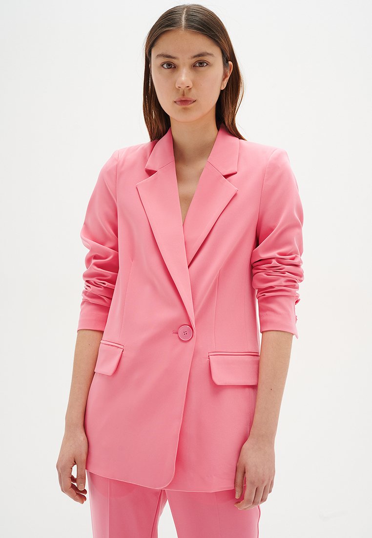 Junge Frau mit langen braunen Haaren, die ein pinkfarbenes, maßgeschneidertes Blazer und passende Hosen trägt, steht vor einem schlichten weißen Hintergrund.