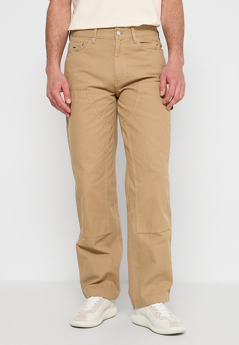 Mann, der beigen gerade geschnittenen Hose mit verstärkten Kniepatches und weißen Sneakers trägt, steht vor einem einfarbigen Hintergrund.