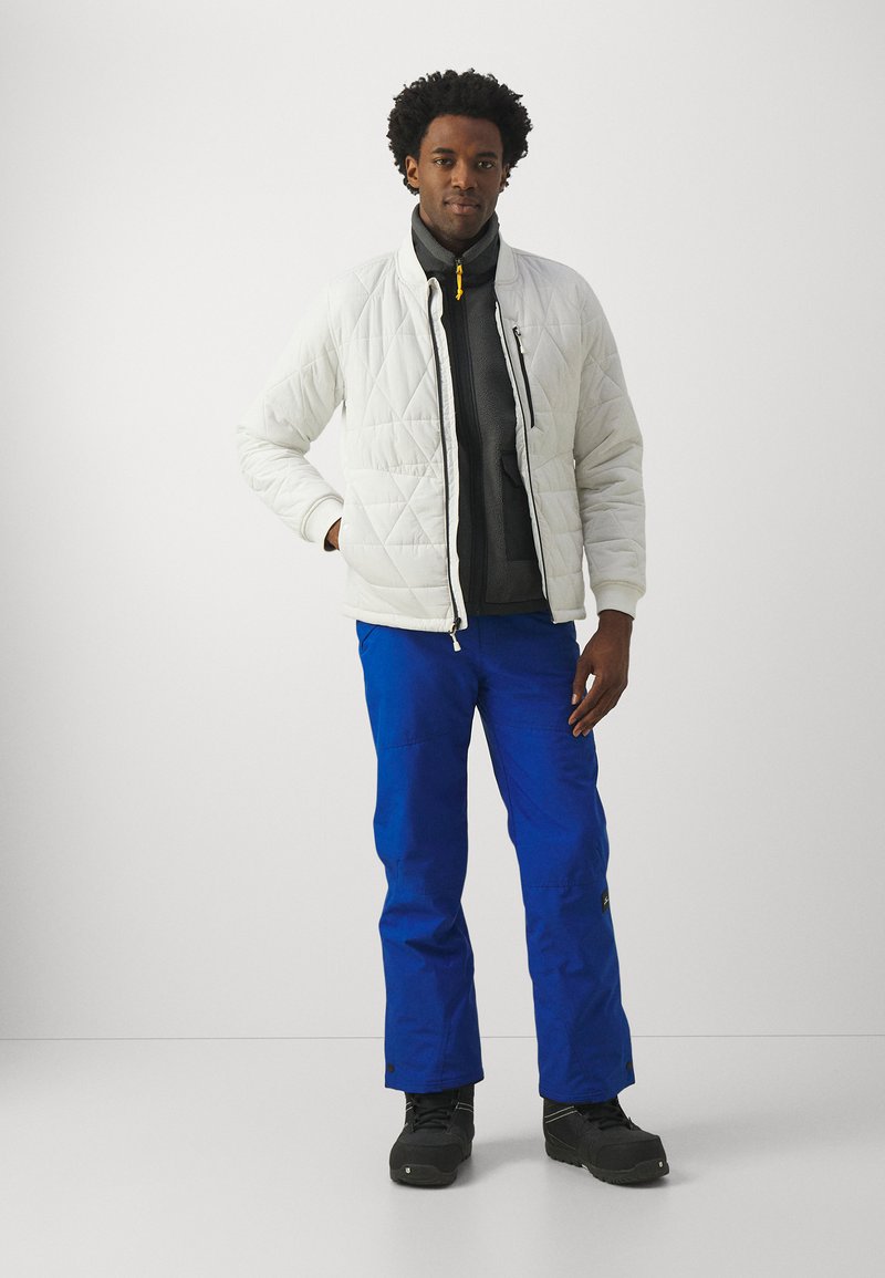 Mann in einer weißen Steppjacke, dunkelgrauem Reißverschluss-Fleece, leuchtend blauen Hosen und schwarzen Schuhen, der vor einem schlichten hellen Hintergrund steht.