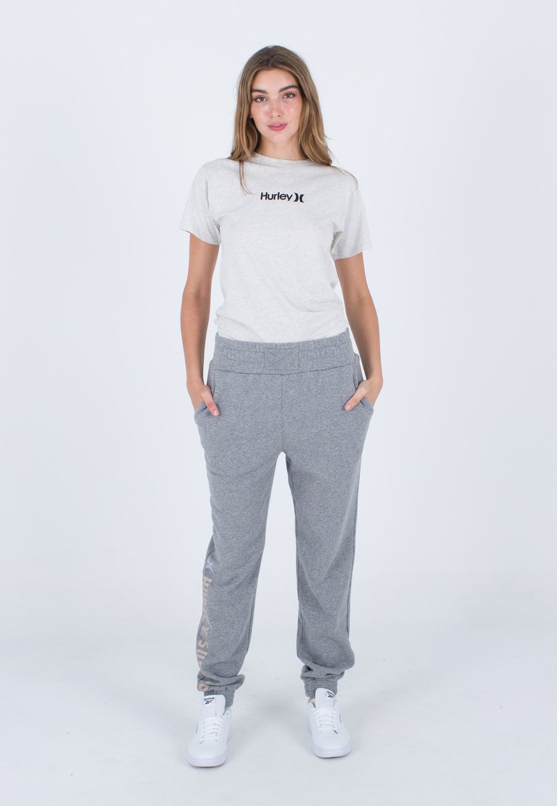 Graues T-Shirt mit kurzen Ärmeln und schwarzem "Hurley"-Logo, kombiniert mit grauen Sweatpants mit strukturiertem Bund und seitlichen Taschen. Weiße Sneaker.