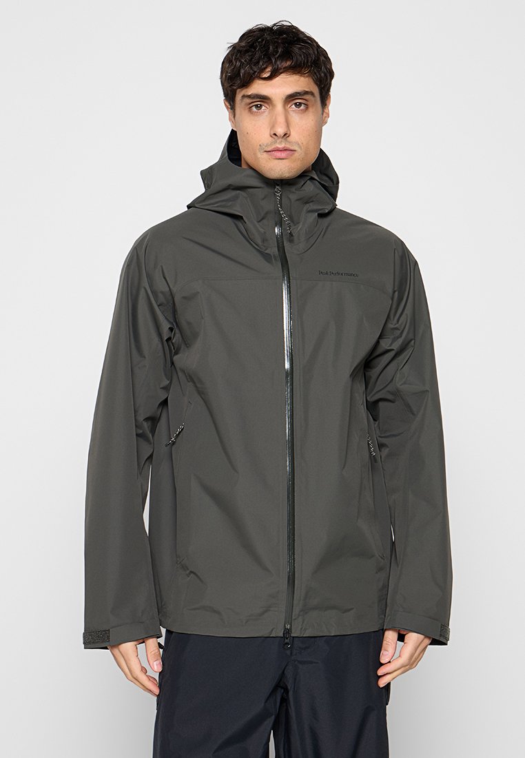 Mann trägt eine dunkelgraue wasserdichte Jacke mit Kapuze, Frontreißverschluss und seitlichen Reißverschlusstaschen, steht vor einem schlichten hellen Hintergrund.