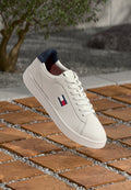 Weiße Ledersneaker mit einem marineblauen Absatz, einem rot-blauen Emblem und weißen Schnürsenkeln, platziert aufHolzfliesen mit Kiesakzenten.