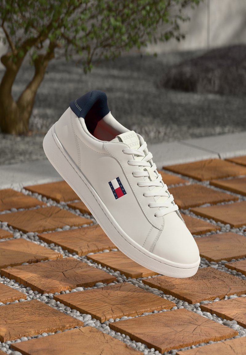 Weiße Ledersneaker mit einem marineblauen Absatz, einem rot-blauen Emblem und weißen Schnürsenkeln, platziert aufHolzfliesen mit Kiesakzenten.