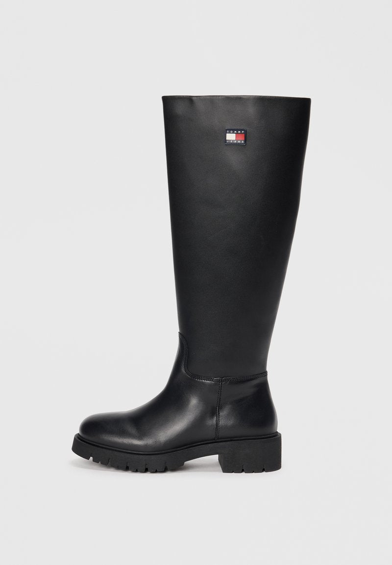 Schwarze knielange Stiefel aus glattem Leder mit einer strukturierten Gummisohle, versehen mit einem kleinen Logoschild in Rot, Weiß und Blau.