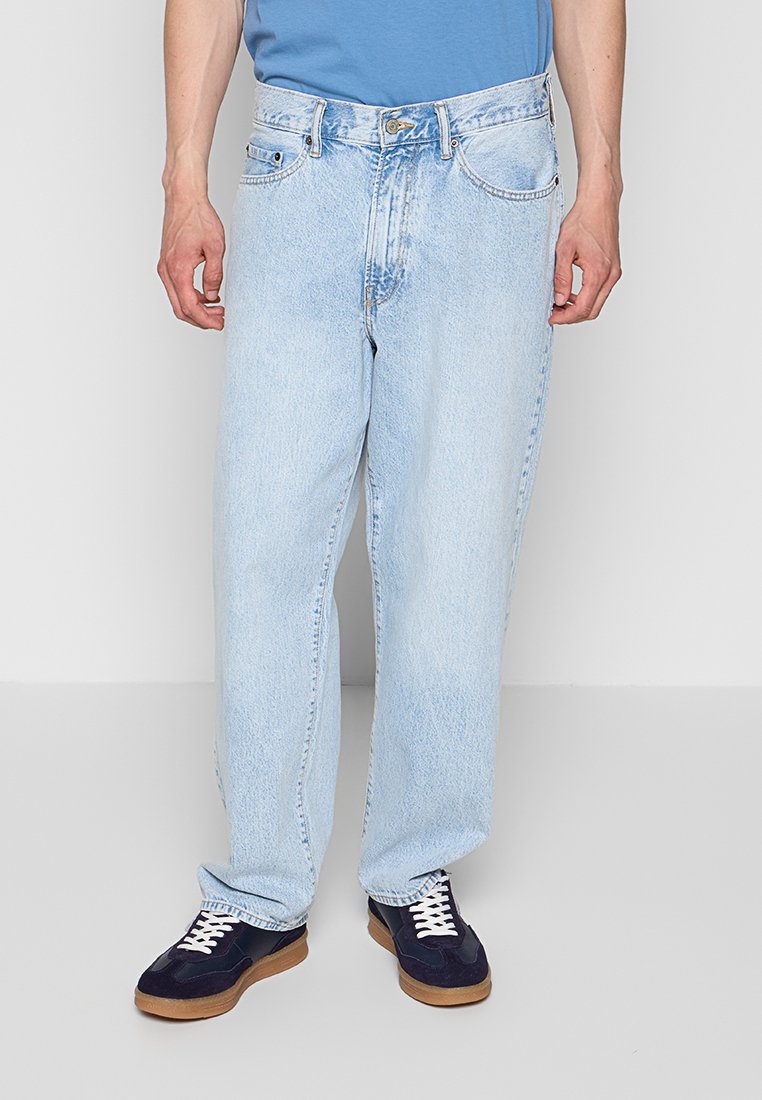 Mann trägt hellblaue, locker sitzende Jeans, blaues Hemd und schwarze Sneakers mit weißen Schnürsenkeln, steht vor schlichtem Hintergrund.