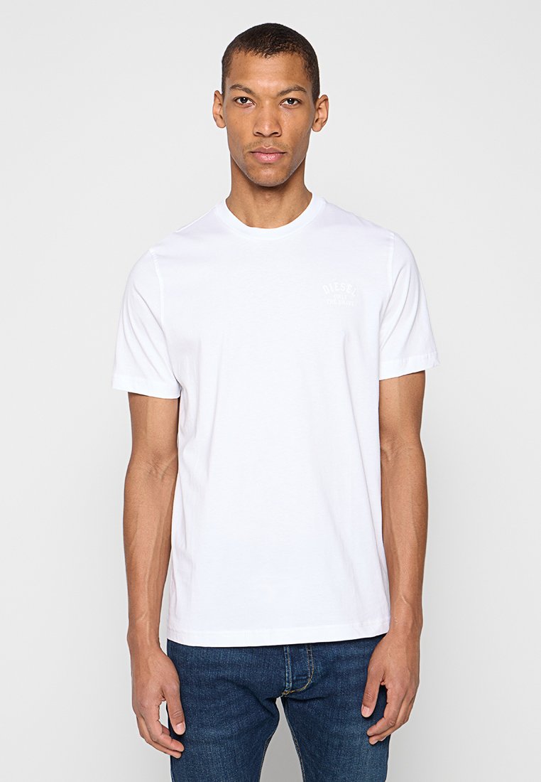 Weißes Baumwoll-T-Shirt mit Rundhalsausschnitt, kurzen Ärmeln und dezentem Markenaufdruck auf der linken Brust. Kombiniert mit dunkelblauer Jeans.