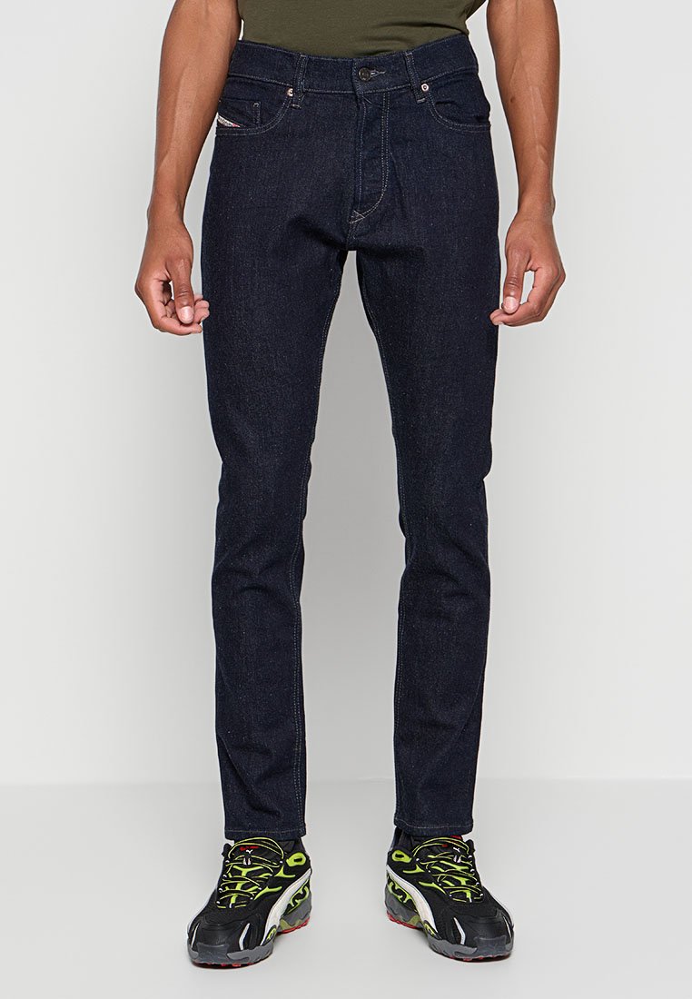 Dunkelblaue Jeans im Slim Fit mit fünf Taschen, design mit kontrastierenden Nähten. Kombiniert mit schwarzen und grünen Sportschuhen.
