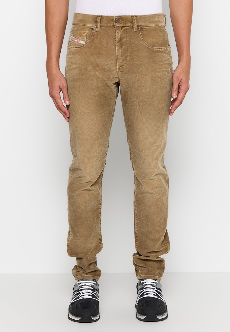 Beige Cord-Hosen mit schmalem Schnitt, Fronttaschen und einem kleinen Logostick. Weiches Material mit subtiler Rippstruktur, kombiniert mit schwarzen Sportschuhen.