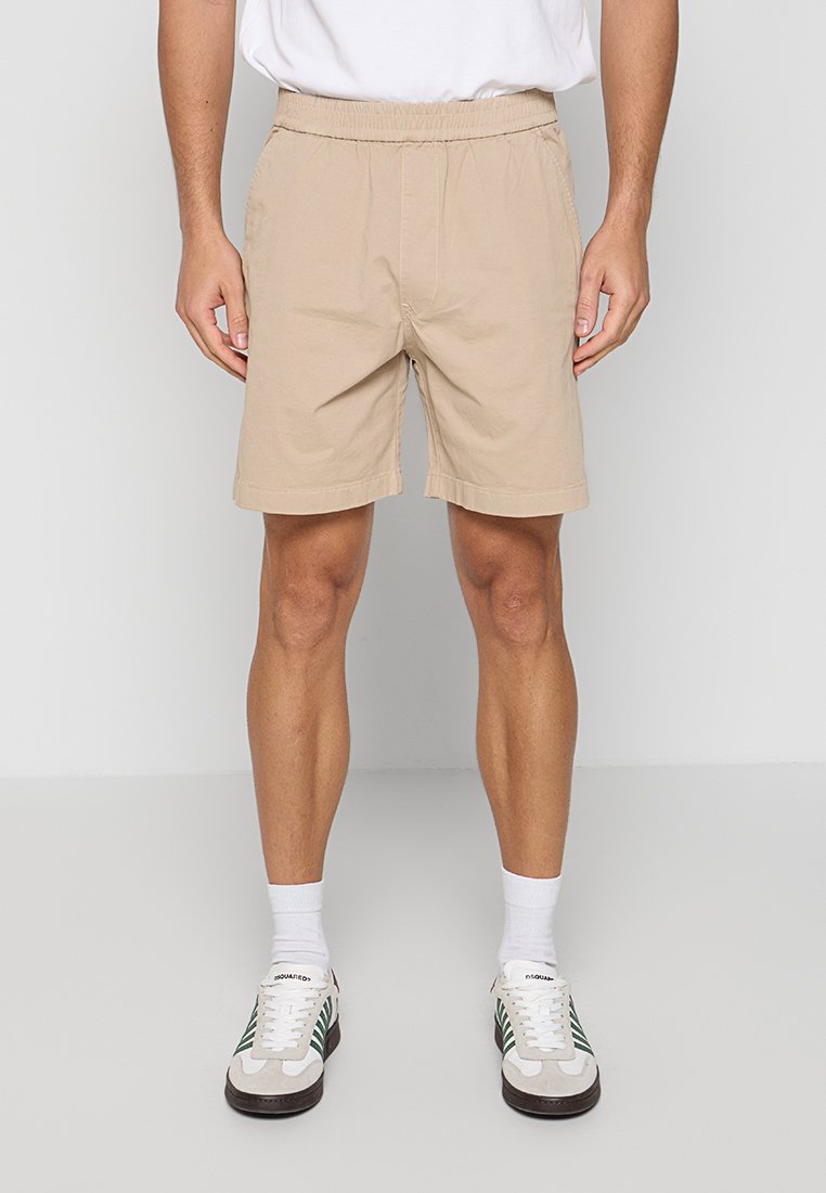 Mann trägt beige Shorts mit elastischem Bund, weiße Socken und weiße Sneaker mit grünen Streifen, steht vor einem einfarbigen hellen Hintergrund.