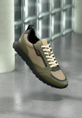 Olivgrüner und beiger Sneaker mit schwarzer Sohle und cremefarbenen Schnürsenkeln, mit rotem "HUGO"-Logo an der Seite, vor einem Hintergrund aus Glasbausteinen präsentiert.