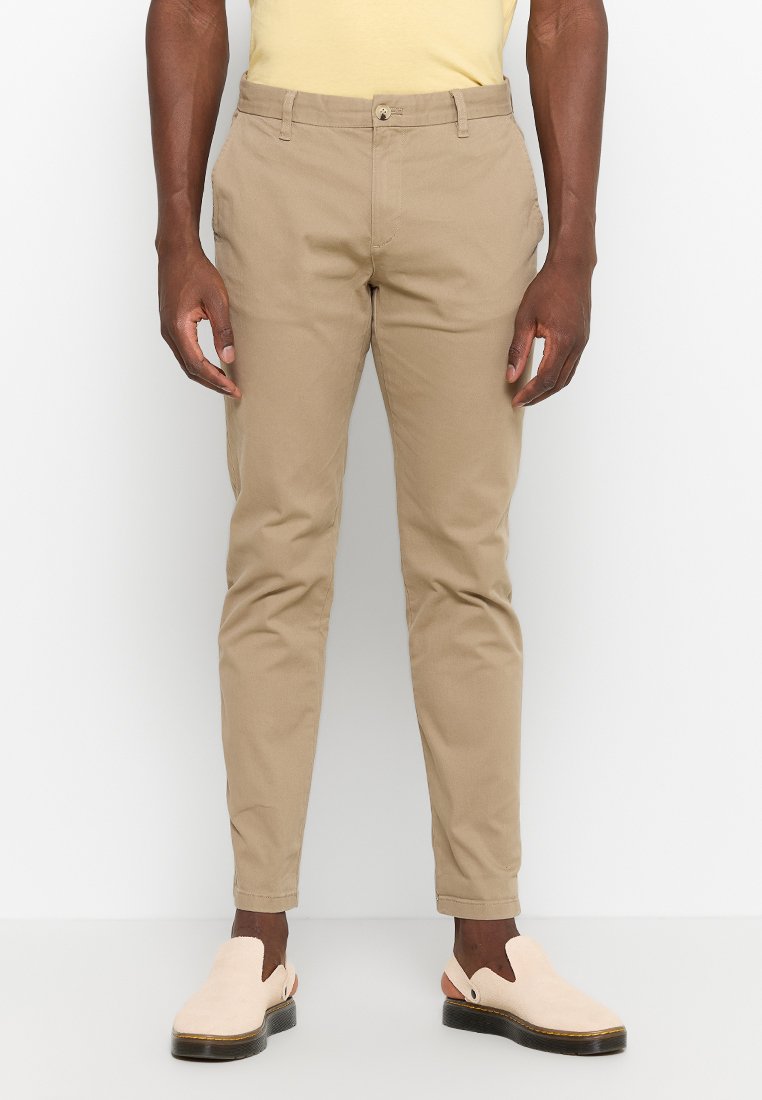Beige Chinos mit schmalem Schnitt, aus Baumwolle. Mit einem Knopfverschluss, Vorderaschen und umgeschlagenen Säumen. Kombiniert mit hellen Slippern.