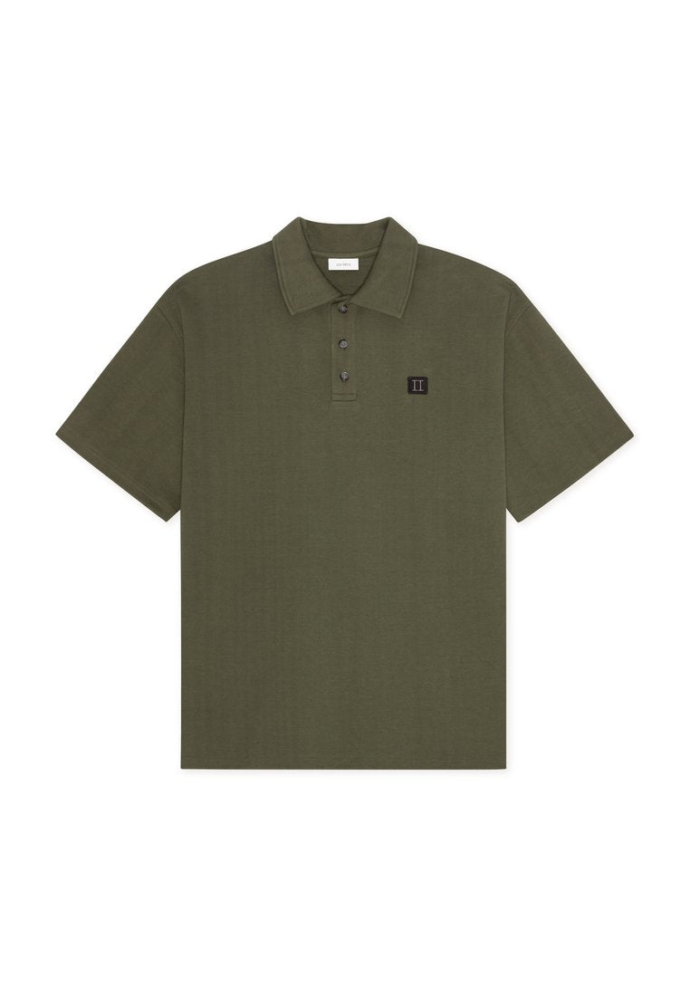 Olivgrünes Poloshirt mit kurzen Ärmeln, drei Knöpfen und kleinem schwarzem quadratischem Patch mit der römischen Zahl II auf der Brust.