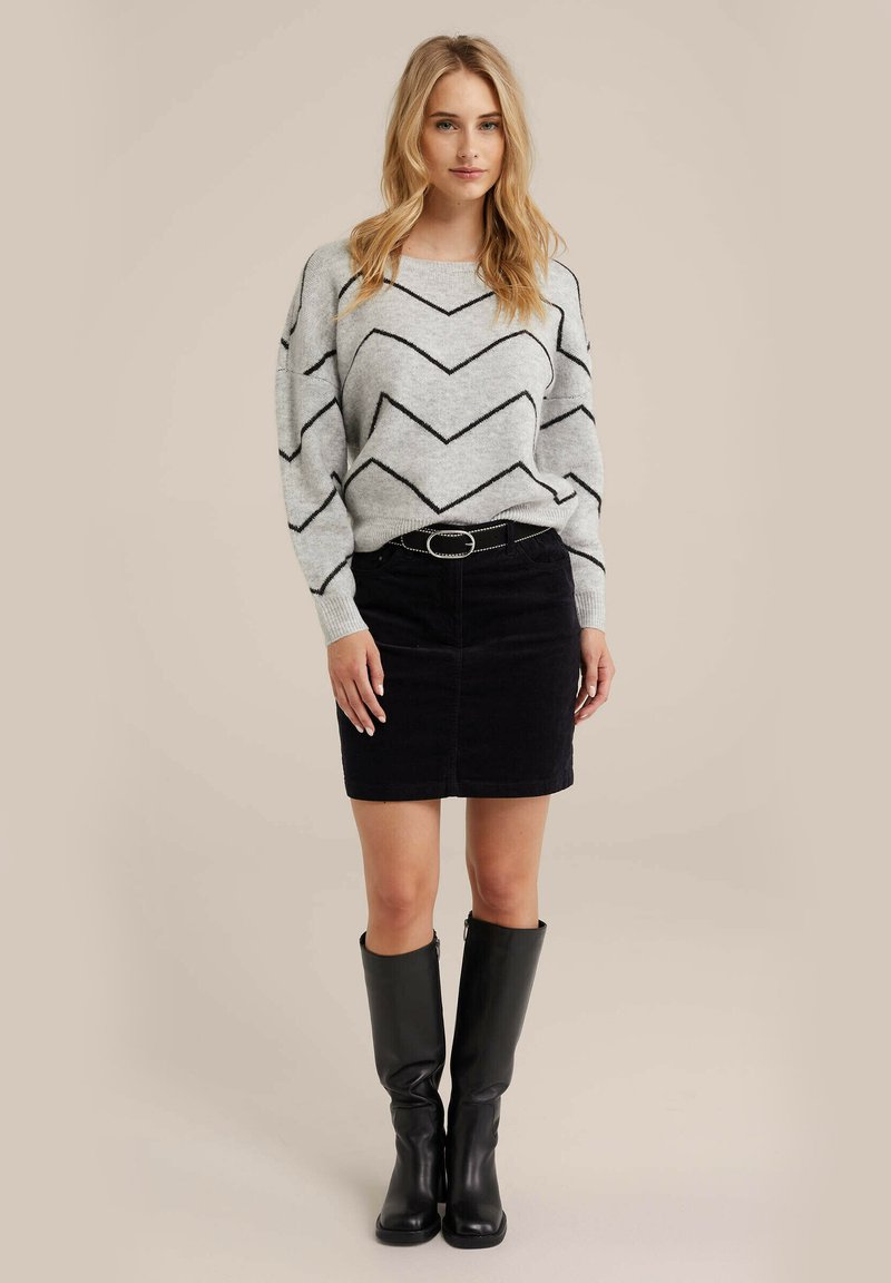 Grauer Pullover mit schwarzem Zickzackmuster, schwarzer Cordrock und knielangen schwarzen Stiefeln, ausgestattet mit einem Gürtel mit einer silbernen Schnalle.