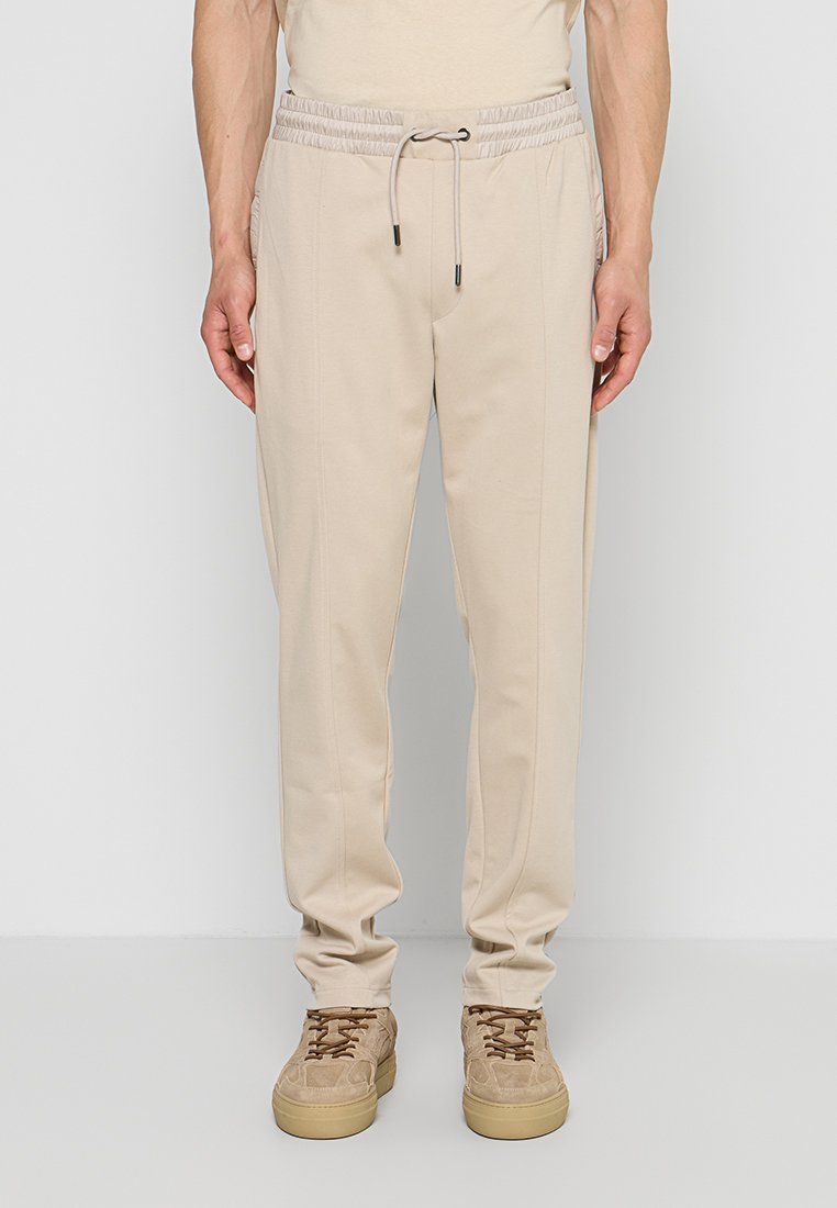 Beige Freizeithose mit elastischem Tunnelzug am Bund, kombiniert mit braunen Wildledersneakern, von der Taille bis zu den Füßen auf neutralem Hintergrund abgebildet.